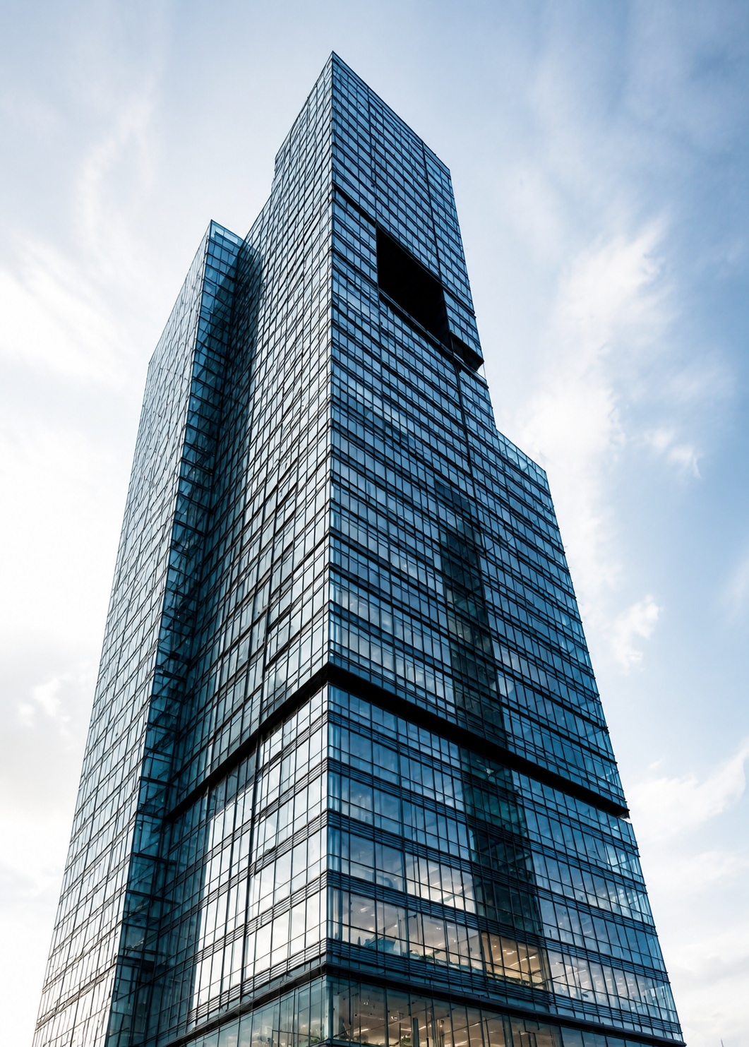 A single glass tower photographed from its base, leaning back into the sky, with a stepped setback partway up and a recessed dark opening near the crown.